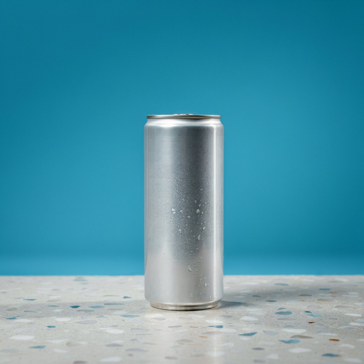 A slim, unbranded sparkling-water can of brushed aluminum, beaded with condensation, stands on a pale terrazzo surface against a seamless electric-blue background; broad overhead diffusion and a crisp side strip create clean highlights, with no lettering or graphics. — Ideogram v4.5