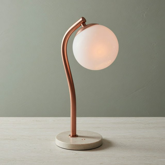 Commercial product photograph of an unbranded sculptural desk lamp with a frosted opal glass globe and brushed copper stem, standing on a pale travertine surface against a muted sage background, illuminated by soft overhead diffusion and a delicate warm rim light. — Grok Imagine Image Quality