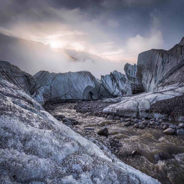 Beyond a braided glacier-fed stream, a steep moraine valley opens beneath clearing dawn snow flurries; gritty ice grains and water-polished stones sharpen the near bank, while layered ridges fade into cloud. — Model 9