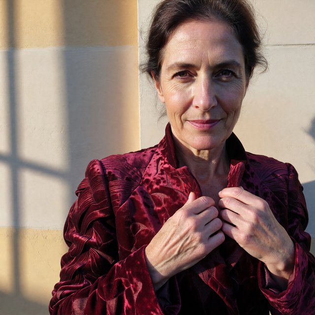 Fashion portrait of an adult woman in her early 50s wearing a sculptural burgundy velvet jacket, her hands lightly gathering its lapel. A wry, composed expression meets the camera against a warm ivory plaster backdrop, lit by soft late-afternoon window glow. — ImagineArt 1.5 Pro