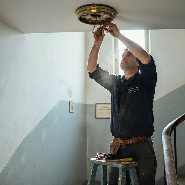 Candid documentary photograph of an adult building caretaker changing a ceiling light in a quiet apartment stairwell, pale afternoon window light, 35mm lens with moderate depth of field, plaster dust drifting from the fixture. — Ideogram v4