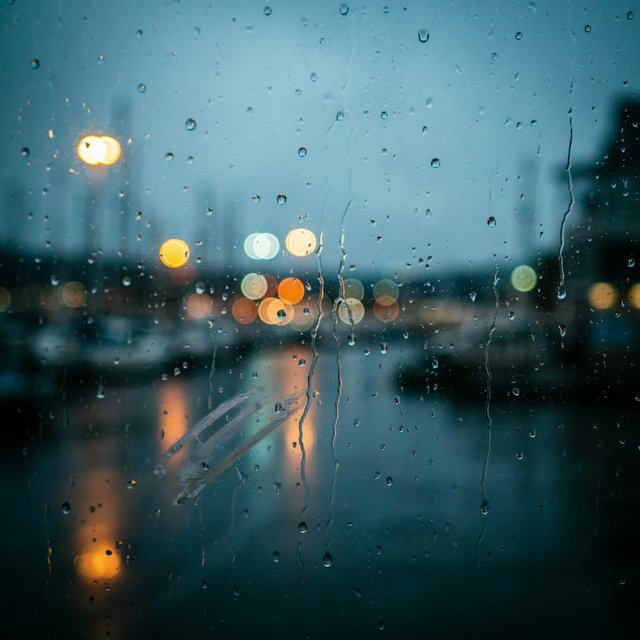Raindrops crawl down a bus shelter pane, warping blurred harbor lights beyond. A handheld evening street snapshot in cool sodium glow, shot at 50mm with shallow depth of field, catches tiny water trails and a faint wipe mark. — Ideogram v4.5