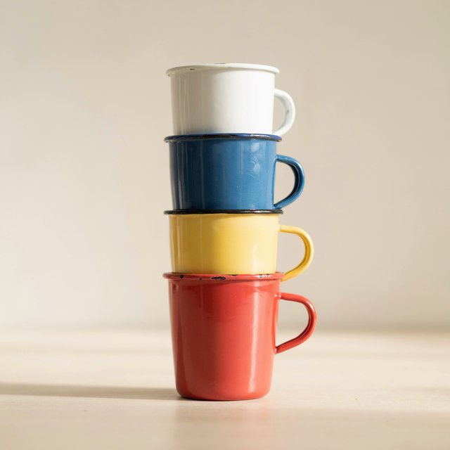Exactly four enamel camping cups form a vertical tower on a pale wooden shelf, bottom to top: red, yellow, blue, white; all handles face right. Soft morning light, nothing else. — Ideogram v4.5