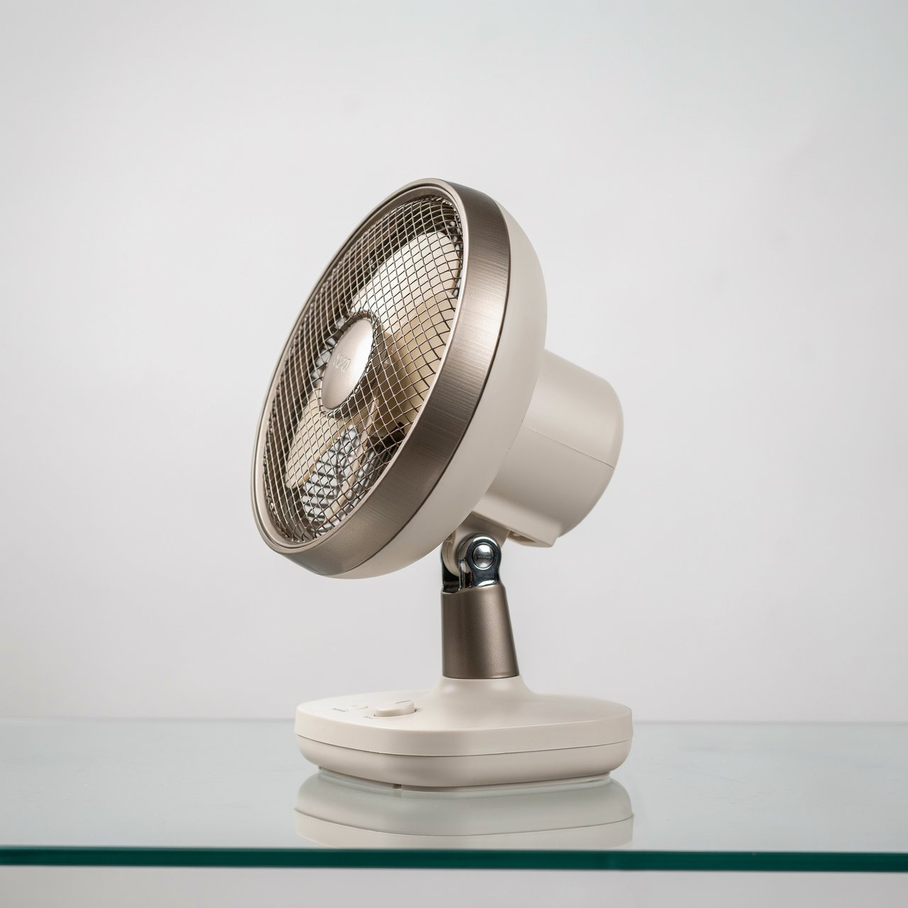 Commercial product photograph of an unbranded desktop fan made from brushed aluminum and matte ivory polymer, resting on a smoked glass surface against a pale gray background, illuminated by broad diffused studio light and a soft rear rim light. — Ideogram v4