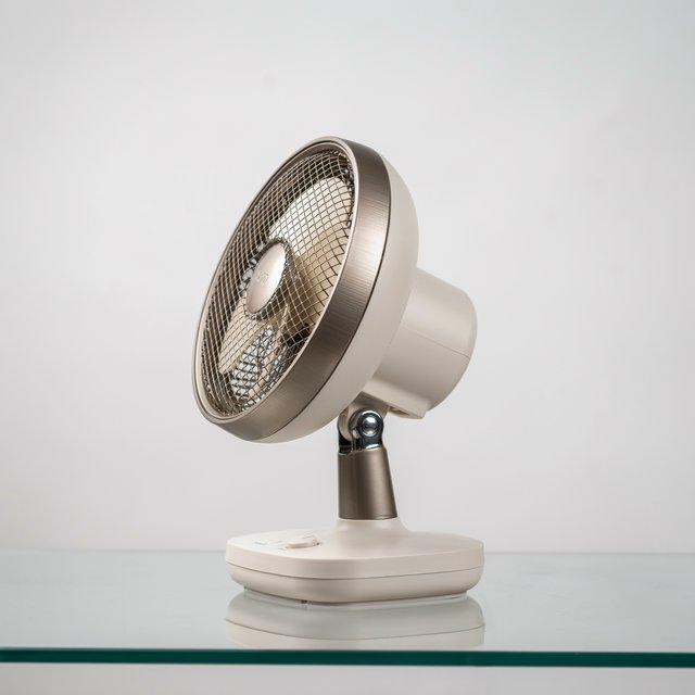 Commercial product photograph of an unbranded desktop fan made from brushed aluminum and matte ivory polymer, resting on a smoked glass surface against a pale gray background, illuminated by broad diffused studio light and a soft rear rim light. — Ideogram v4