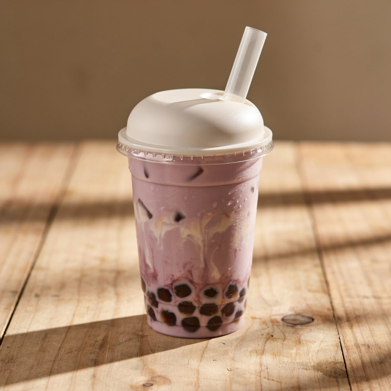 Photorealistic product shot of iced taro bubble tea in a clear frosted plastic cup, condensation beading down curved sides, wide clear straw piercing matte white domed lid. Plump dark brown tapioca pearls settled at the base, swirled creamy purple tea with soft marbled cream streaks, fine ice crystals glinting inside. Resting on weathered light oak table, soft golden hour side lighting casting gentle long shadows, warm neutral beige background. Shallow depth of field, sharp focus on glass texture and pearl details, professional food photography aesthetic, soft moody warm colour palette with rich lavender and caramel tones. — Ideogram v4.5