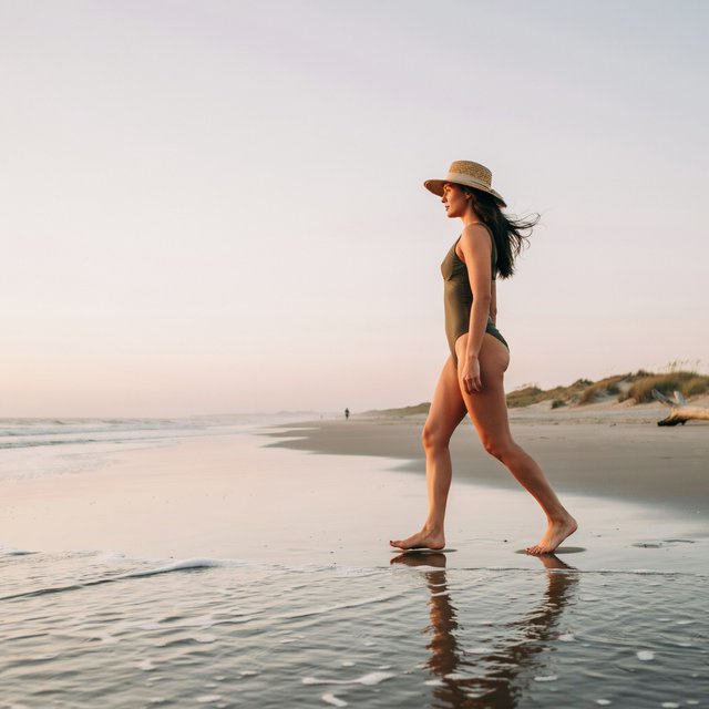 Editorial beach photo of an adult woman in a one-piece swimsuit and a wide straw hat walking along the waterline at sunset, wind in her hair. — Ideogram v4