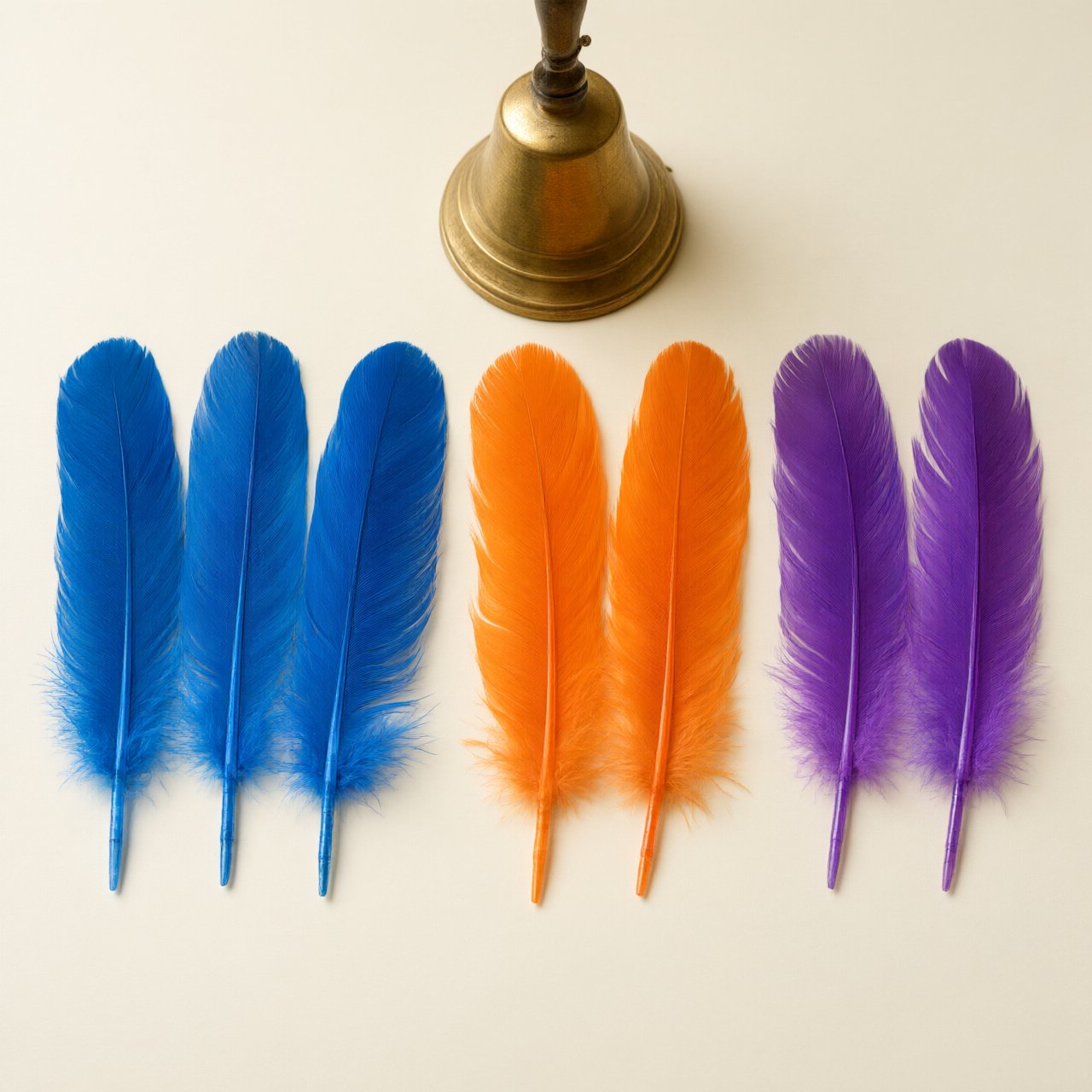 Exactly seven feathers lie on a cream tabletop: three blue on the left, two orange in the center, and two purple on the right. A brass bell sits above the orange pair, nothing else. — Qwen Image 3.0