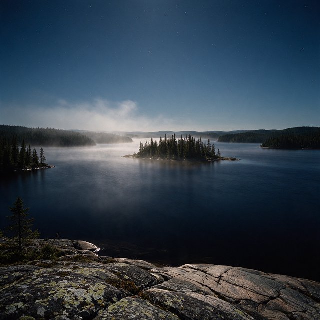 An expansive boreal lake beneath a clear, cold midnight sky, pale moonlight catching thin mist above dark water; spruce-covered islands recede toward distant ridges, while lichen and cracked granite sharpen the near shoreline. — Seedream 5.0 Pro