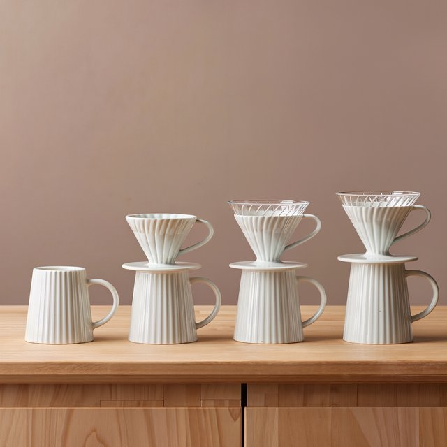 From directly overhead, an unbranded pour-over coffee set with ribbed porcelain and clear borosilicate components arranged in sequence on pale oak; warm taupe background, broad diffused softbox light and gentle side fill shape this commercial product image, no lettering. — Model 9