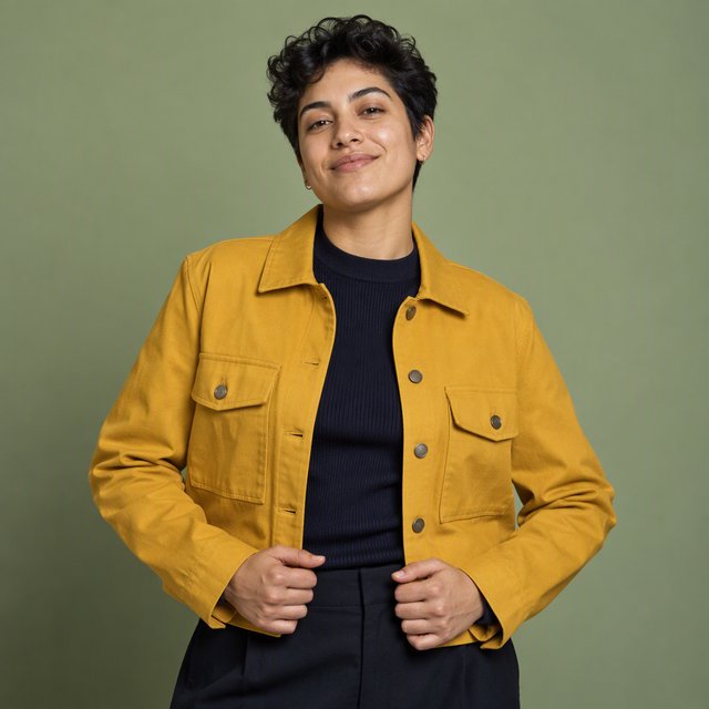 Fashion portrait of an adult nonbinary person in their late twenties wearing a structured saffron-yellow canvas jacket over a fine-gauge navy merino top, hands resting on the jacket hem. Their bright, self-assured expression contrasts with a softly lit sage-green backdrop. — GPT Image 2.5 Sunburst
