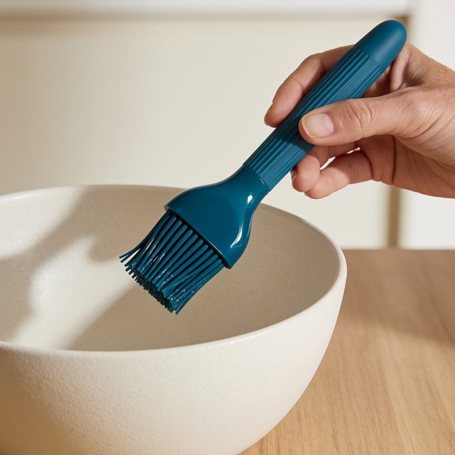 Across a honed maple counter, an adult hand guides an unbranded silicone pastry brush over a ceramic bowl; its ribbed teal handle catches diffused key light and warm bounce against a cream kitchen background, face cropped out, no lettering, commercial product image. — Krea 2 Medium