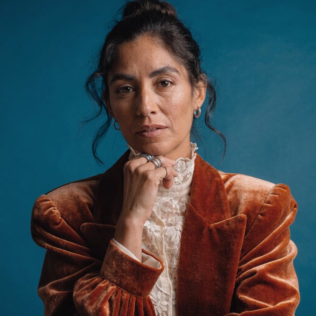 Portrait of an adult woman in her 30s wearing a rust-colored velvet jacket over an ivory blouse, one hand resting beneath her chin, thoughtful expression, deep teal studio backdrop, soft side lighting and gentle cheek highlights. — Recraft v4