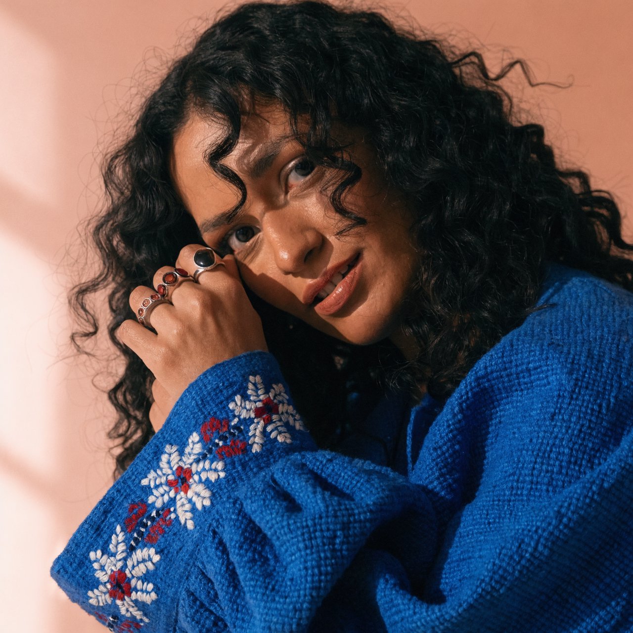 Fashion portrait of an adult Latina woman in her late 20s wearing a handwoven cobalt-blue cotton jacket, one hand touching its embroidered cuff, curious half-smile, against a pale peach studio backdrop, soft diffused window light shaping her face. — Recraft v4 Pro