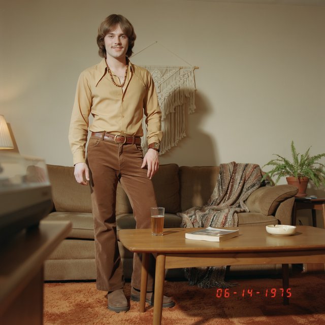 Create an image of a man in the quintessential style of 1975. Style his hair, clothing, accessories, and surroundings to reflect the distinctive look of the mid-1970s—featuring bold silhouettes, eye-catching accessories, layered details, and characteristic colors and textures. Give the image the feel of an authentic 1975 photograph, complete with analog film grain, faded colors, direct flash lighting, and a slight soft-focus effect. Add a period-accurate, red-orange date stamp in a 1970s style in one of the bottom corners. No recognizable posters. No modern objects or text. — Ideogram v4