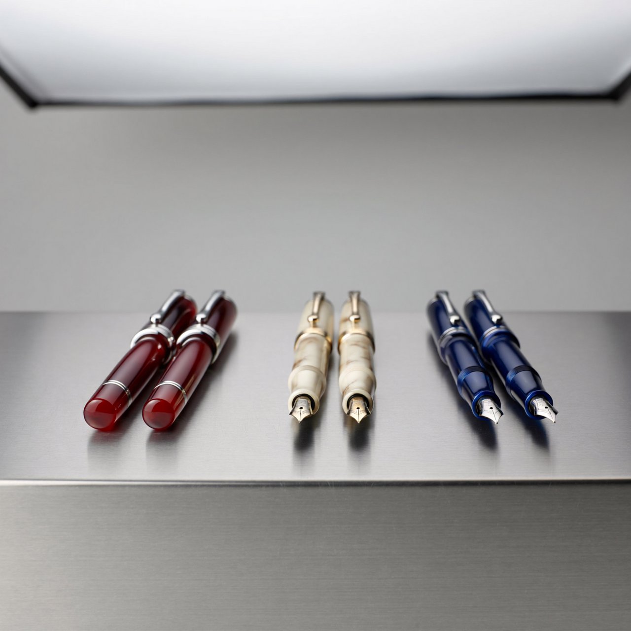 Across a brushed steel counter, exactly six fountain pens rest in three groups: two crimson pens on the left, two ivory pens in the center, and two cobalt pens on the right; nothing else. — ImagineArt 2