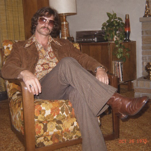 Create an image of a man in the quintessential style of 1975. Style his hair, clothing, accessories, and surroundings to reflect the distinctive look of the mid-1970s—featuring bold silhouettes, eye-catching accessories, layered details, and characteristic colors and textures. Give the image the feel of an authentic 1975 photograph, complete with analog film grain, faded colors, direct flash lighting, and a slight soft-focus effect. Add a period-accurate, red-orange date stamp in a 1970s style in one of the bottom corners. No recognizable posters. No modern objects or text. — GPT Image 2.5 Flare