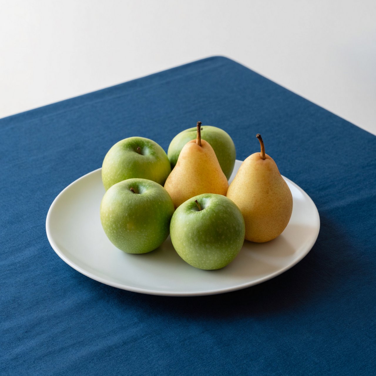 Exactly three green apples and two yellow pears on a white plate, the plate on a blue tablecloth, nothing else on the table. — ImagineArt 2