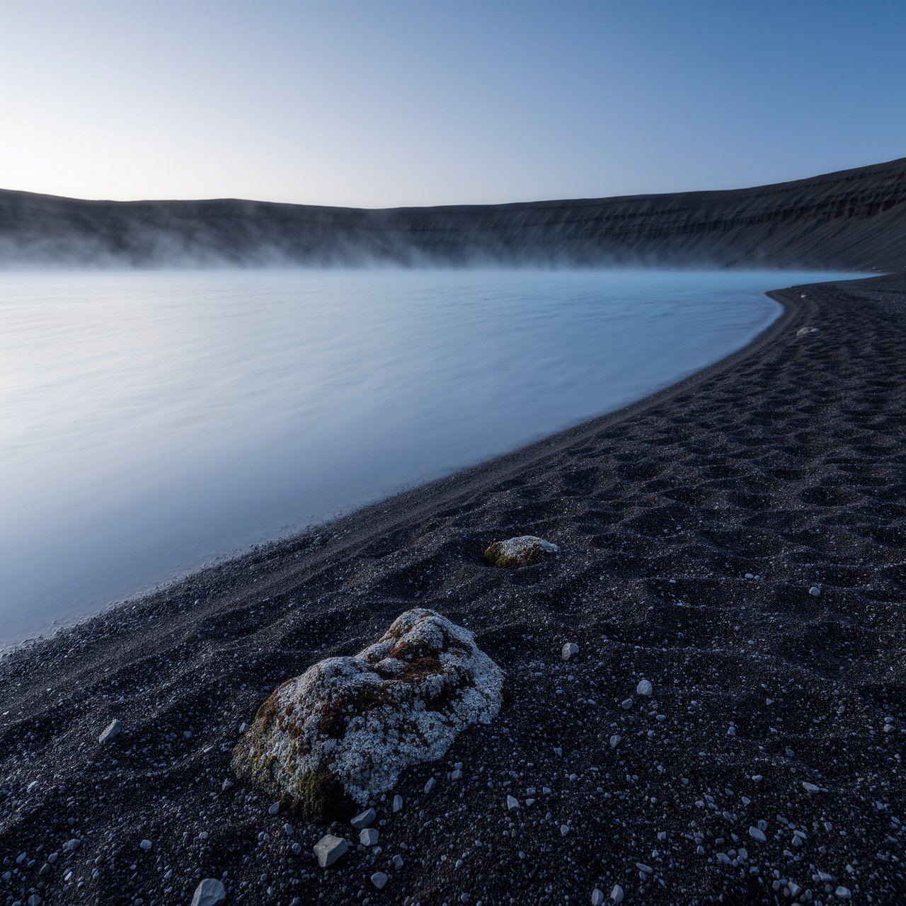 At first light, a rain-cleared volcanic caldera lake lies beneath thinning fog; pumice grains and damp lichen sharpen the near shore, while dark cinder slopes step back toward a distant rim. — Kling v3 Omni