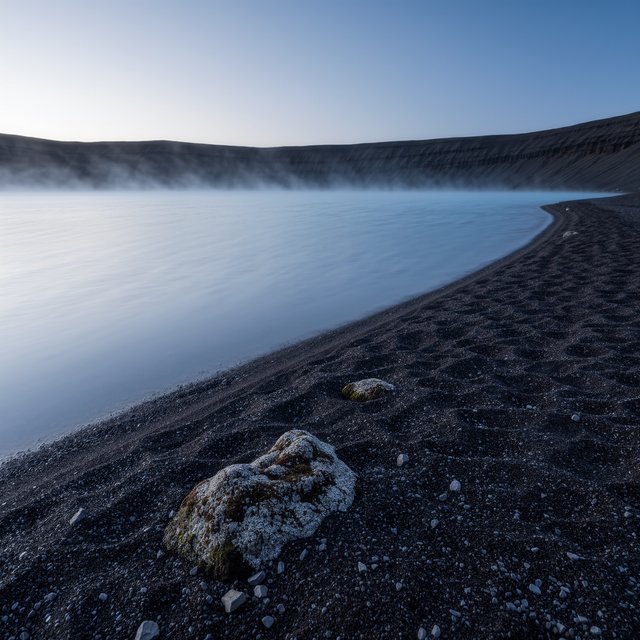 At first light, a rain-cleared volcanic caldera lake lies beneath thinning fog; pumice grains and damp lichen sharpen the near shore, while dark cinder slopes step back toward a distant rim. — Kling v3 Omni