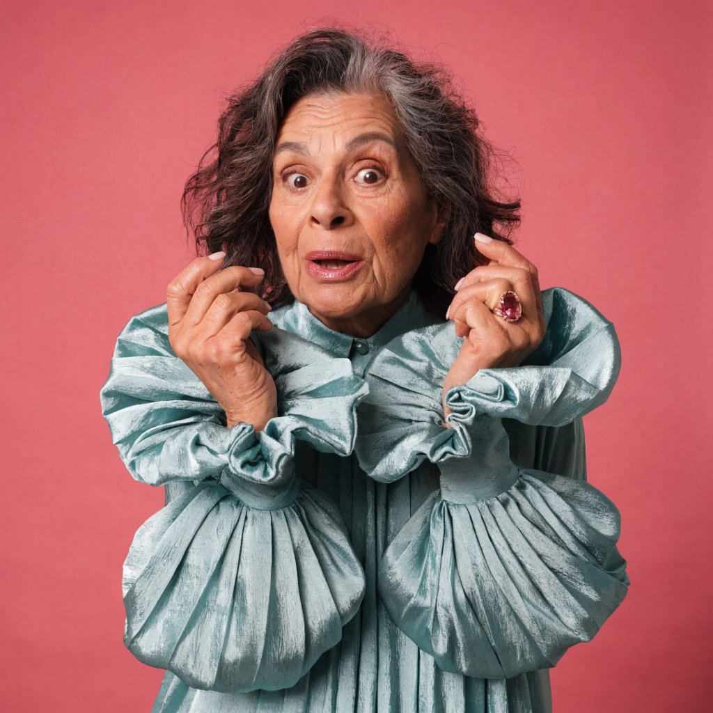 Portrait photograph of an adult woman in her early 60s wearing a softly pleated sea-green silk blouse, hands brushing its gathered sleeve fabric. Her amused, open expression stands against a matte rose backdrop, illuminated by broad, gentle studio light. — Recraft v4