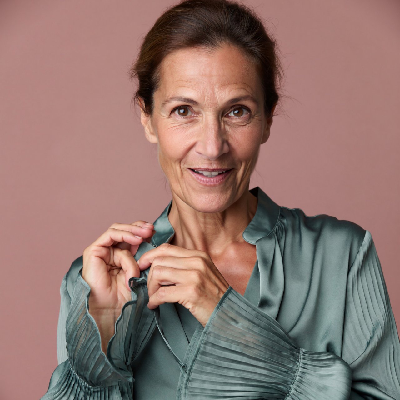 Portrait photograph of an adult woman in her early 60s wearing a softly pleated sea-green silk blouse, hands brushing its gathered sleeve fabric. Her amused, open expression stands against a matte rose backdrop, illuminated by broad, gentle studio light. — ImagineArt 2