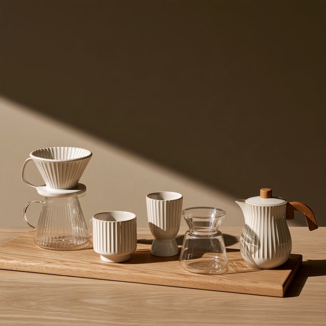 From directly overhead, an unbranded pour-over coffee set with ribbed porcelain and clear borosilicate components arranged in sequence on pale oak; warm taupe background, broad diffused softbox light and gentle side fill shape this commercial product image, no lettering. — Model 13