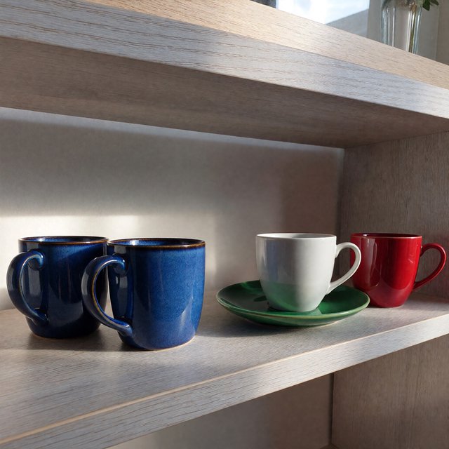 Exactly four ceramic mugs sit on a pale wooden shelf: two blue mugs on the left, one white mug in the center, and one red mug on the right. A green saucer is beneath the center mug, nothing else. — ImagineArt 1.5 Pro