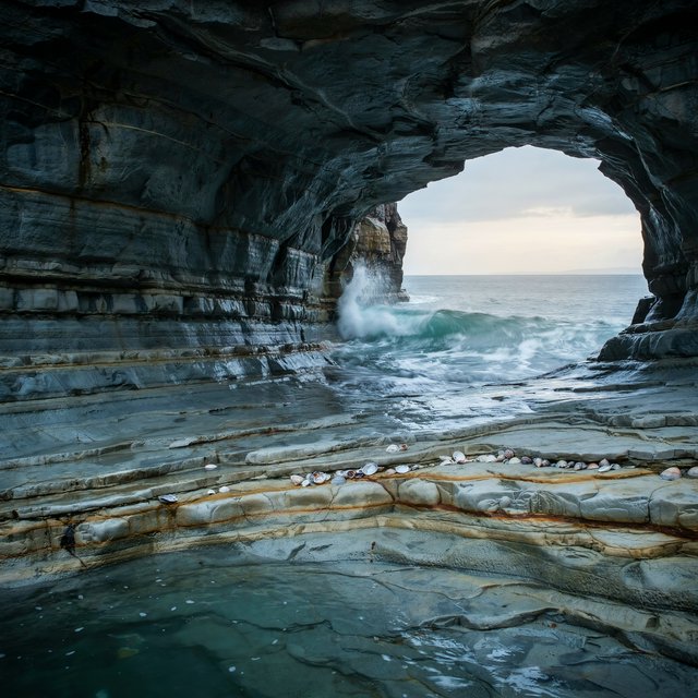 Inside a blue-grey sea cave, incoming swells curl over ribbed limestone beneath a clearing rain squall in late afternoon; wet mineral bands and shell fragments sharpen the entrance ledge, while the cavern mouth frames hazy open water. — Ideogram v4.5