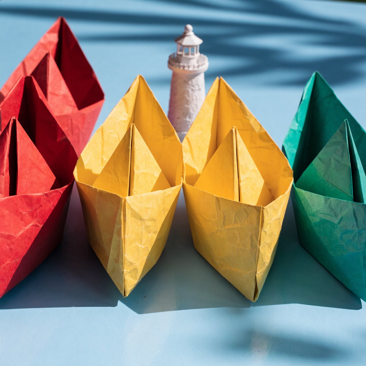 Exactly six folded paper boats sit on a pale blue tabletop: two red boats on the left, two yellow boats in the center, and two green boats on the right. A small white lighthouse figurine stands behind the center pair, nothing else. — Recraft v4 Pro