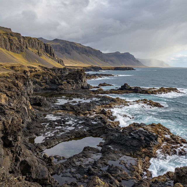 A rugged volcanic coastline under clearing storm clouds at late afternoon, black basalt shelves descending into turquoise surf, sea spray clinging to jagged rocks, tide pools reflecting distant cliffs and layered mountains fading into rain haze. — Qwen Image 3.0 Pro