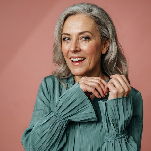 Portrait photograph of an adult woman in her early 60s wearing a softly pleated sea-green silk blouse, hands brushing its gathered sleeve fabric. Her amused, open expression stands against a matte rose backdrop, illuminated by broad, gentle studio light. — Ideogram v4.5