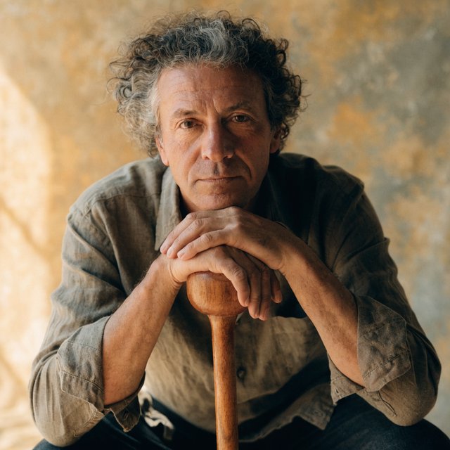 Portrait photograph of an adult man in his 40s with silver-streaked hair, wearing a textured olive linen shirt and rolled sleeves, hands resting on a wooden cane, relaxed expression, ochre plaster backdrop, soft window light from the side. — Recraft v4