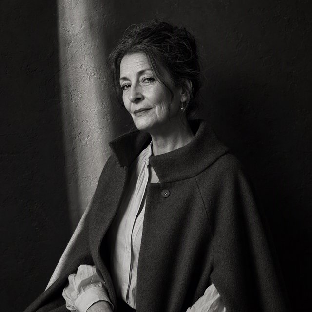 Under a narrow window beam, an adult woman in her late 50s wears a sculpted wool cape over a cotton blouse; her wry expression rests against a charcoal plaster backdrop in a black-and-white portrait, shaped by soft angled light. — GPT Image 2