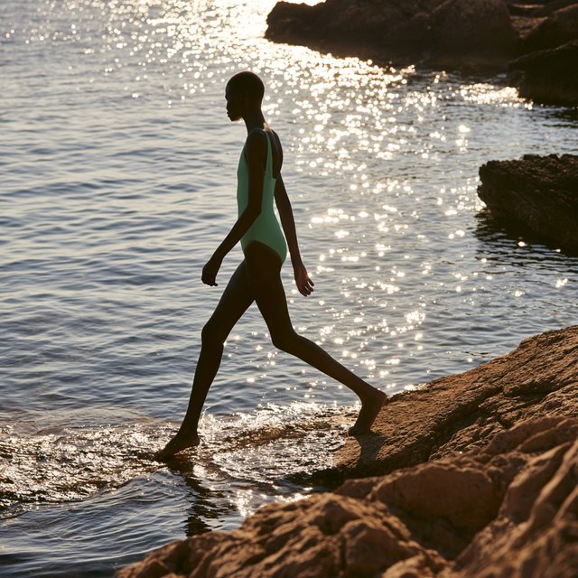 Golden late-afternoon light skims a sheltered cove as an adult woman in a jade one-piece swimsuit is caught mid-step across sunlit rocks, her gaze turned toward the sea; crisp reflections and composed styling bring fashion-magazine polish. — Midjourney V8.2