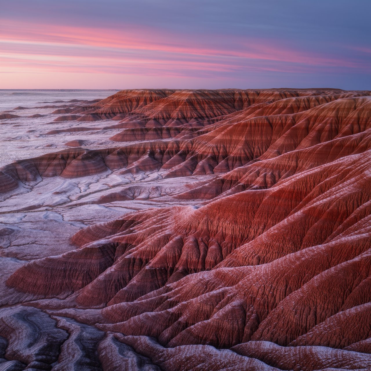 A red-sandstone badlands plateau beneath clearing winter clouds at sunrise, overnight frost silvering the gullies; cracked clay and wind-smoothed rock lead the eye across layered mesas fading into pale distance. — Kling v3 Omni