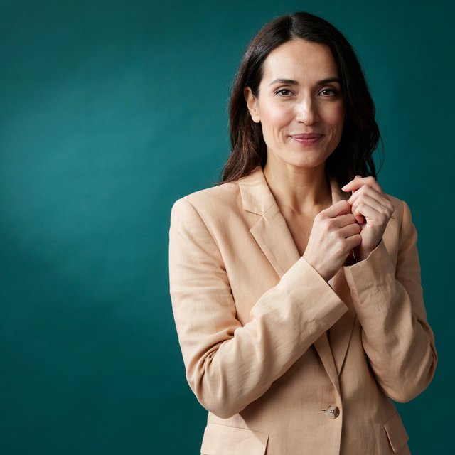 Portrait photograph of an adult woman in her early 30s wearing a pale apricot linen blazer, her hands gently smoothing its cuff. She looks quietly delighted against a deep teal paper backdrop, illuminated by soft, angled studio light. — Ideogram v4.5