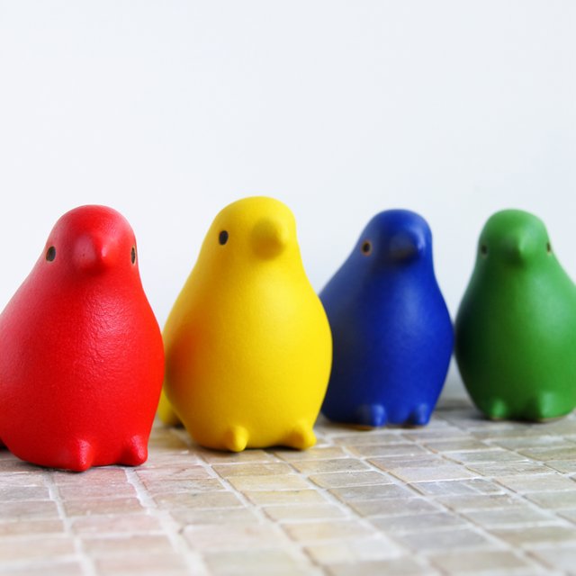 On a sunlit tiled ledge, exactly four ceramic bird figurines form a row: red at far left, yellow at left-center, blue at right-center, and green at far right; all face forward, nothing else. — Krea 2 Large