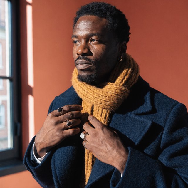 Editorial portrait of an adult Black man in his 30s wearing a deep navy wool overcoat and mustard scarf, fingers adjusting the scarf, thoughtful expression, muted terracotta studio backdrop, crisp cool window light. — Recraft v4 Pro
