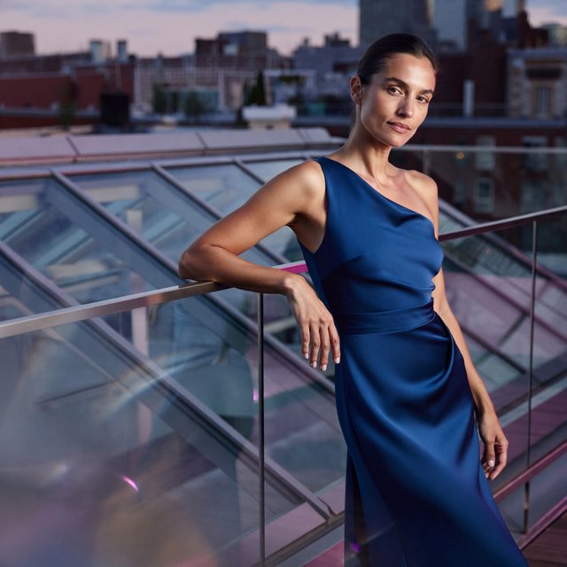 Editorial glamour photograph of one adult woman in a sapphire one-shoulder evening gown, leaning naturally against a rooftop conservatory railing at dusk, flattering lavender light, glass reflections, composed expression, elegant fashion-magazine atmosphere. — ImagineArt 2