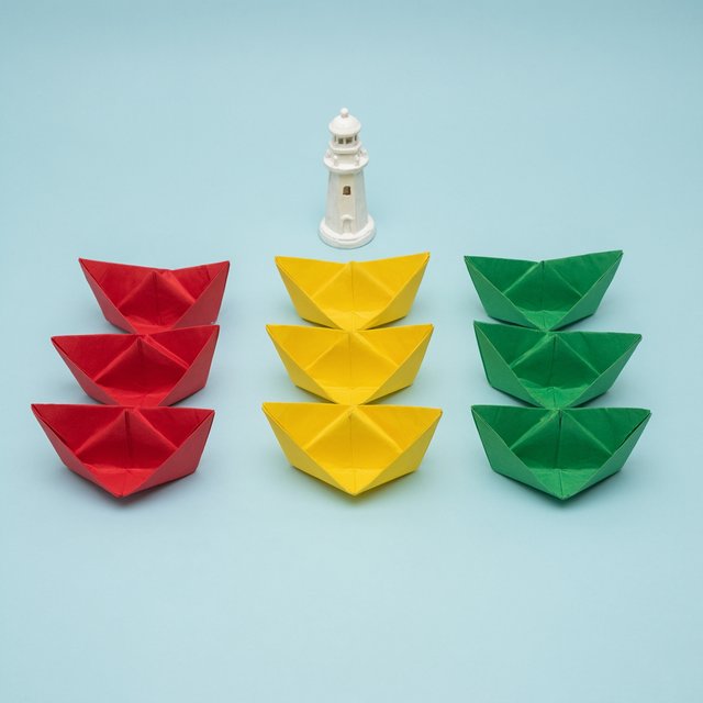 Exactly six folded paper boats sit on a pale blue tabletop: two red boats on the left, two yellow boats in the center, and two green boats on the right. A small white lighthouse figurine stands behind the center pair, nothing else. — Qwen Image 3.0 Pro