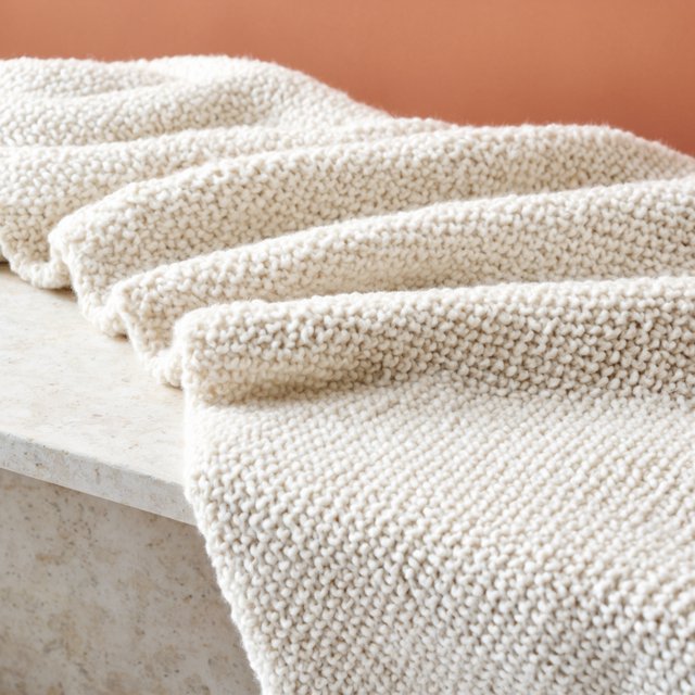 Soft folds of an unbranded chunky-knit wool throw rest on a pale travertine bench; its looped surface catches broad diffused key light and gentle side fill against a muted terracotta studio background, forming a clean commercial product image with no lettering. — Model 11