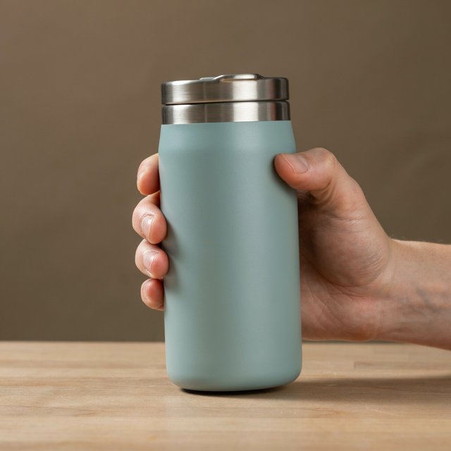 An adult hand lifts an unbranded insulated travel mug, its matte sea-glass polymer body and brushed-steel rim catching softbox highlights above a pale oak counter; warm gray studio backdrop, broad diffused key and cool edge light, no face or lettering. — Ideogram v4.5