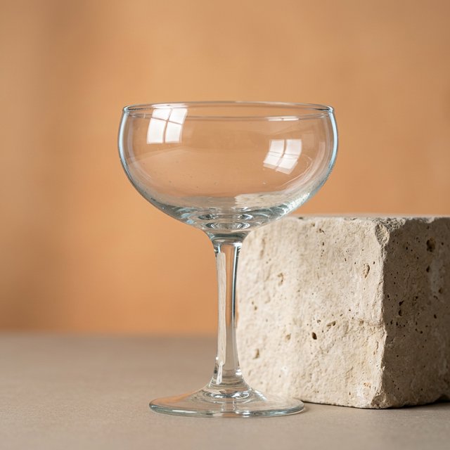 Beside a pale limestone block, an unbranded handblown cocktail coupe in clear soda-lime glass catches crisp window-shaped highlights; its curved bowl bends a muted apricot backdrop under broad diffused key light and narrow cool rim lighting, commercial product image, no lettering. — Nano Banana 2.1