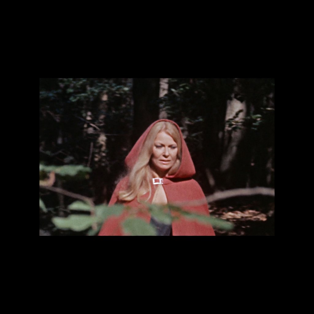 Medium shot at eye level: 1960s vintage film aesthetic. An adult woman with long, blonde hair walks alone through a dark forest. She wears a red hooded cloak, with the hood pulled up and fastened at the neck by a silver buckle. Photorealistic 1960s Technicolor film grain; soft, shallow depth of field. — Krea 2 Medium
