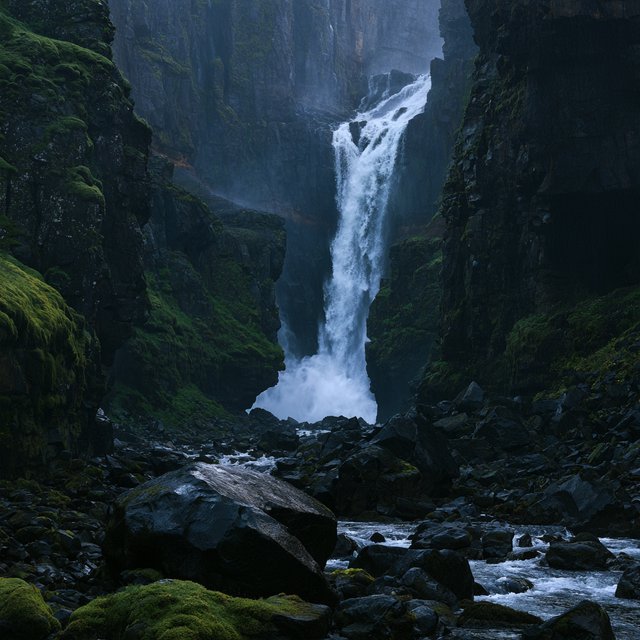 After a passing squall, a waterfall drops through a narrow basalt gorge at dusk, its white plume breaking against dark ledges; rain-beaded moss and sharp foreground stones lead toward cliffs fading behind drifting spray. — Midjourney V8.2