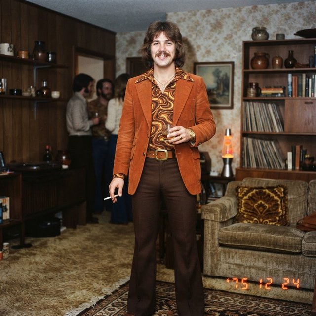Create an image of a man in the quintessential style of 1975. Style his hair, clothing, accessories, and surroundings to reflect the distinctive look of the mid-1970s—featuring bold silhouettes, eye-catching accessories, layered details, and characteristic colors and textures. Give the image the feel of an authentic 1975 photograph, complete with analog film grain, faded colors, direct flash lighting, and a slight soft-focus effect. Add a period-accurate, red-orange date stamp in a 1970s style in one of the bottom corners. No recognizable posters. No modern objects or text. — Nano Banana 2 Lite