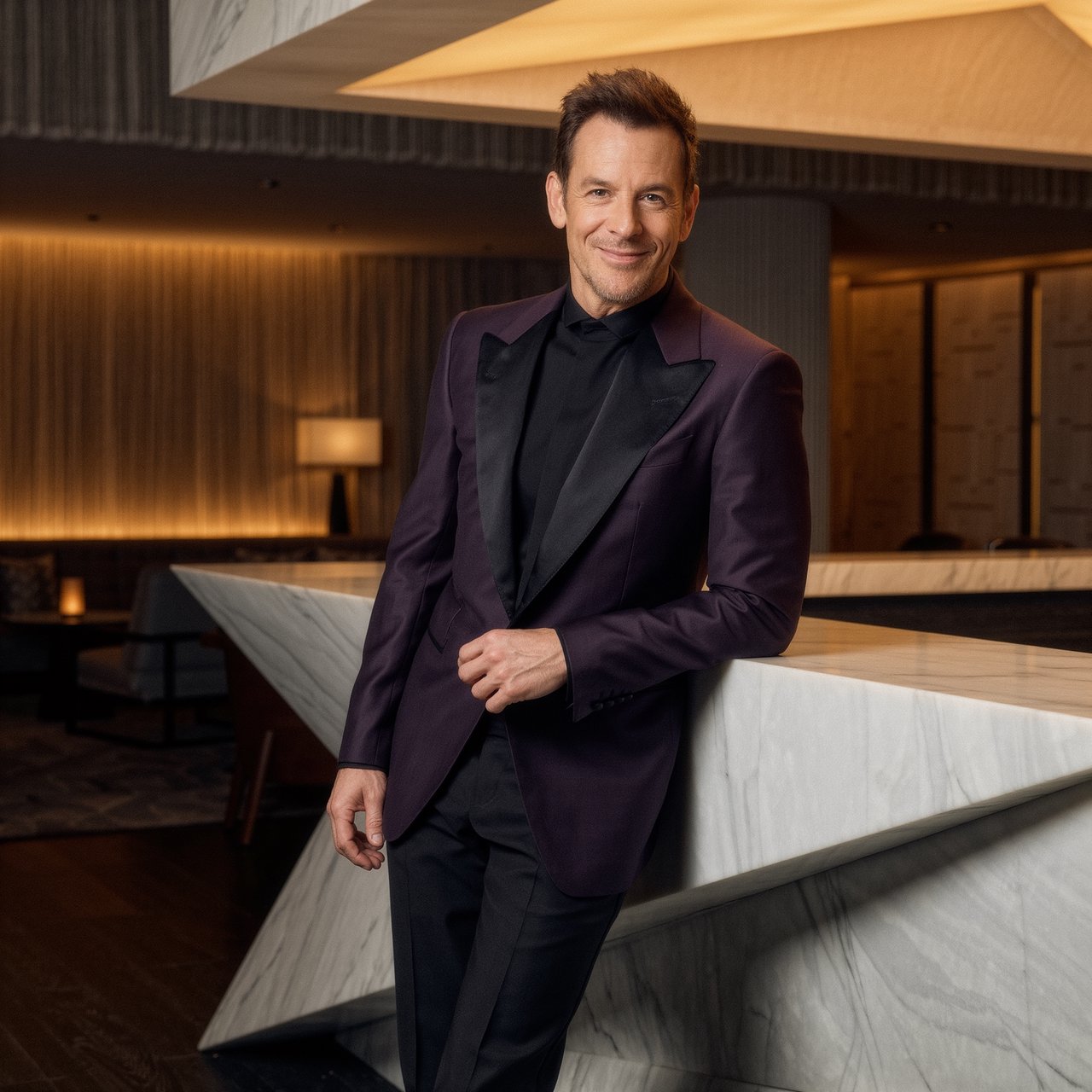 Editorial glamour photograph of an adult man in a midnight-plum dinner jacket and black trousers, standing naturally beside a sculptural marble bar in a quiet hotel lounge, flattering amber light, easy smile, polished editorial elegance. — Kling v3 Omni