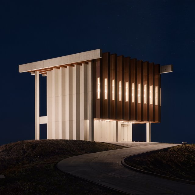 A hilltop observatory in spare contemporary architecture glows against the midnight sky, its pale concrete walls, bronze panels, and narrow windows lit from within; viewed from the access road, crisp edges and credible structural supports. — Kling v3