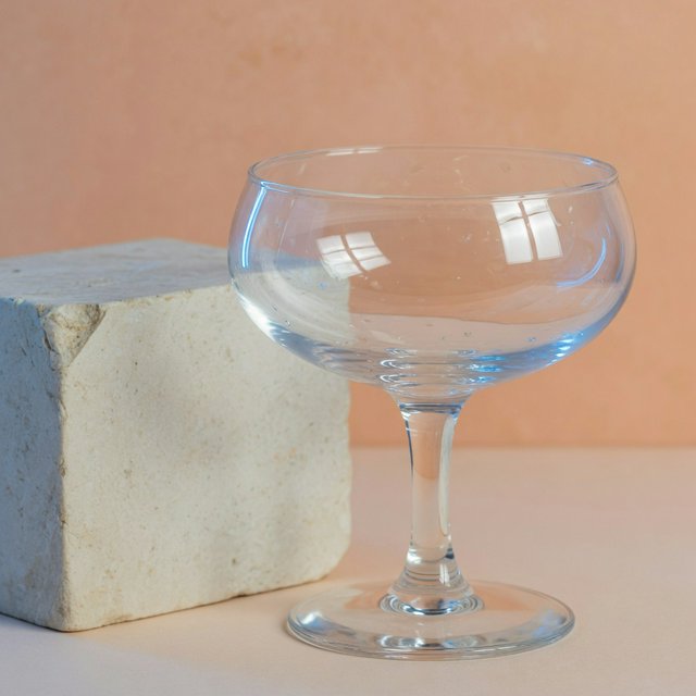 Beside a pale limestone block, an unbranded handblown cocktail coupe in clear soda-lime glass catches crisp window-shaped highlights; its curved bowl bends a muted apricot backdrop under broad diffused key light and narrow cool rim lighting, commercial product image, no lettering. — Grok Imagine Image Quality
